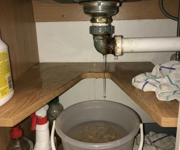 Fuite d'eau importante sous un évier de cuisine, montrant l'eau s'écoulant par le raccord de siphon dans un seau. Plombier sur Bruxelles pour réparation d'urgence.